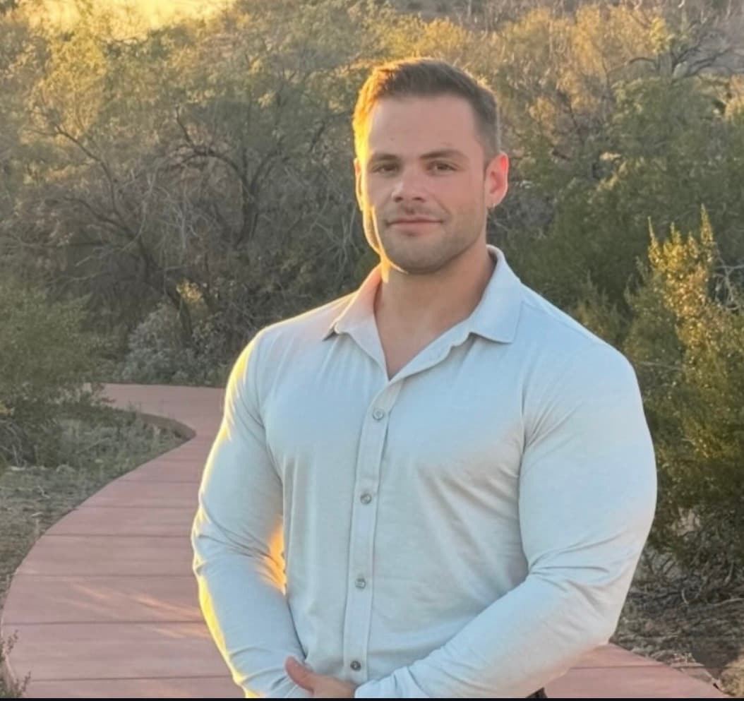 Muscular man in a light button-down shirt standing on an outdoor path during sunset.