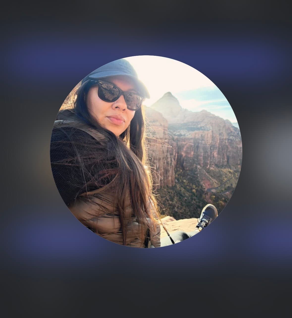 Woman in sunglasses and a cap sits overlooking a vast, sunlit canyon and mountain range.