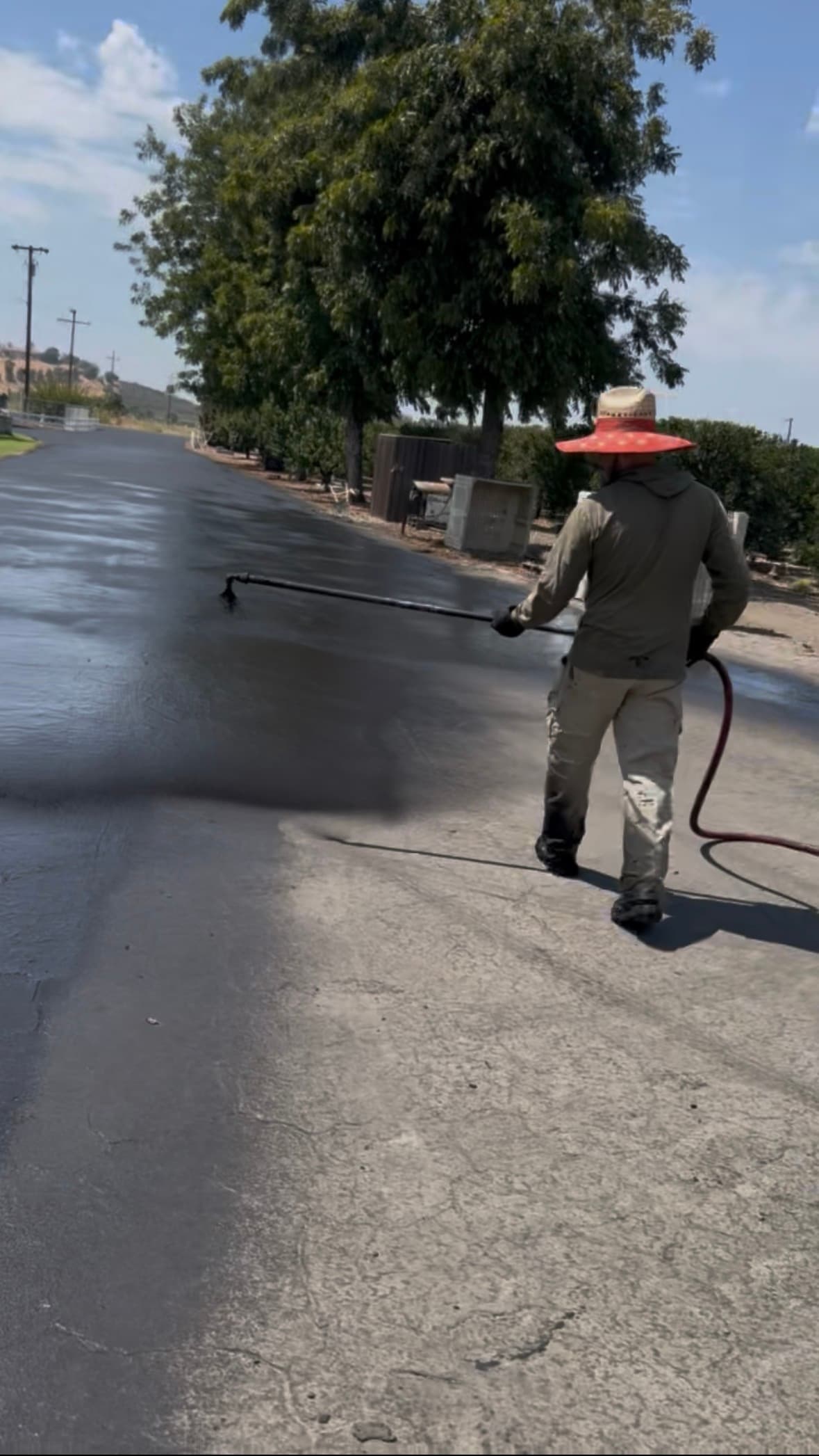 Man in a straw hat sprays black asphalt sealer onto a long driveway outdoors.
