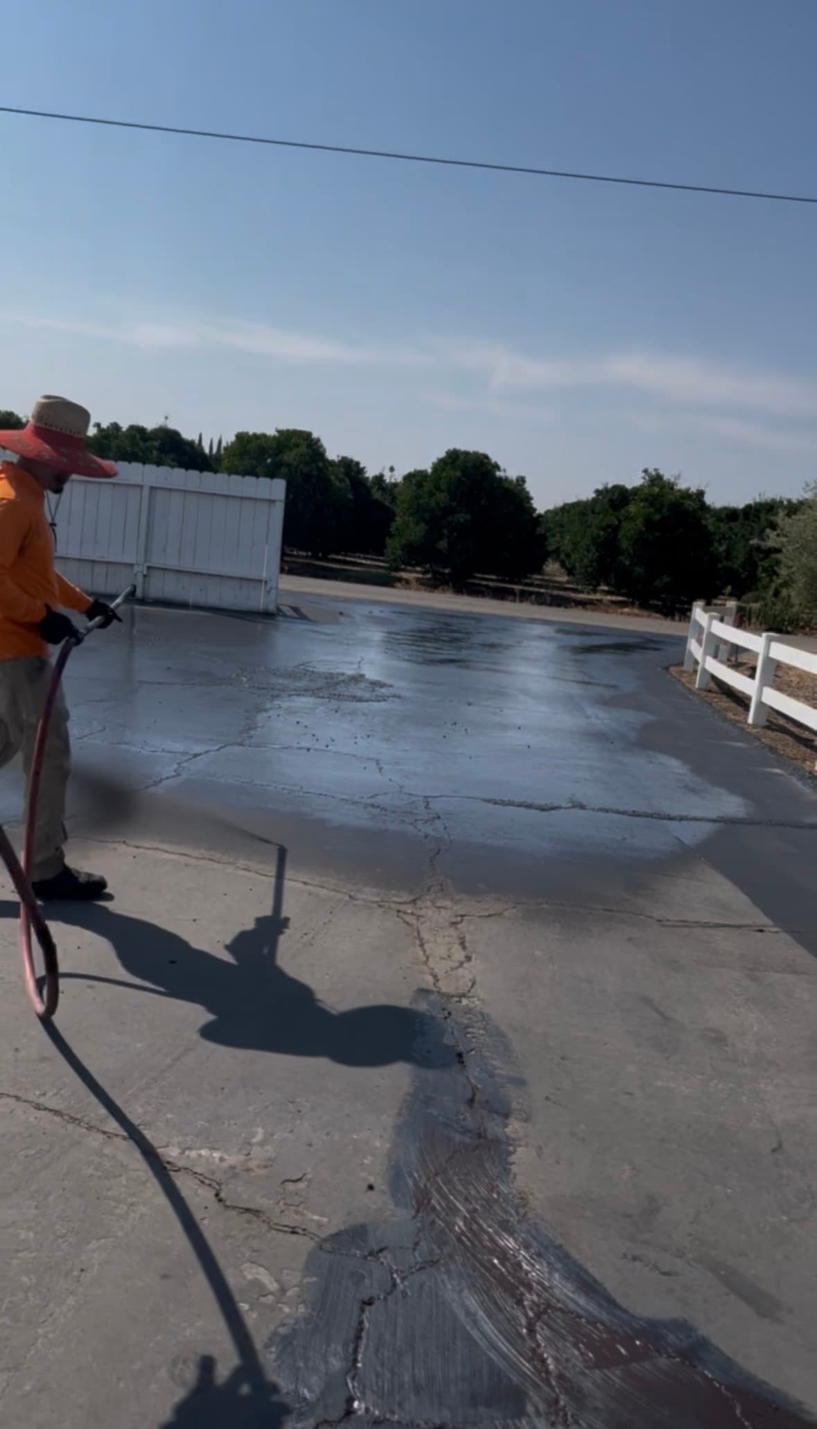 Worker in an orange shirt sprays black sealant onto a cracked asphalt driveway outdoors.