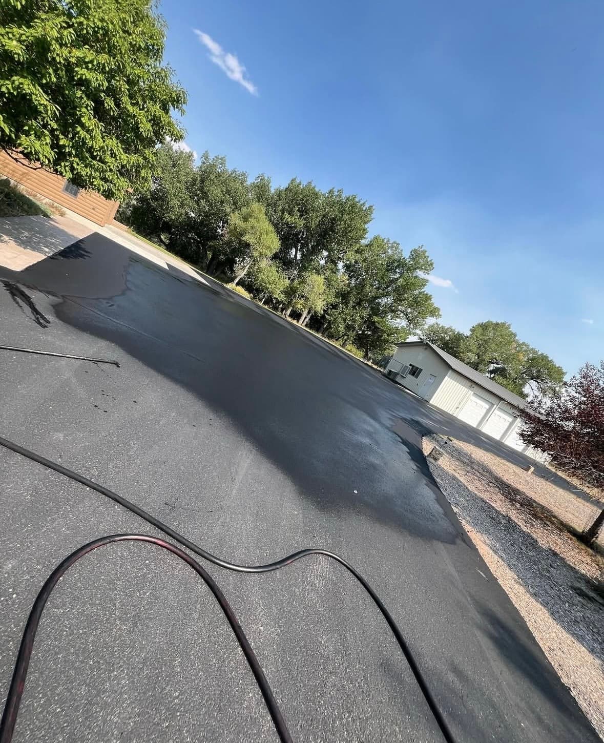 Freshly sealed black asphalt driveway with a hose, trees, and a white garage building.