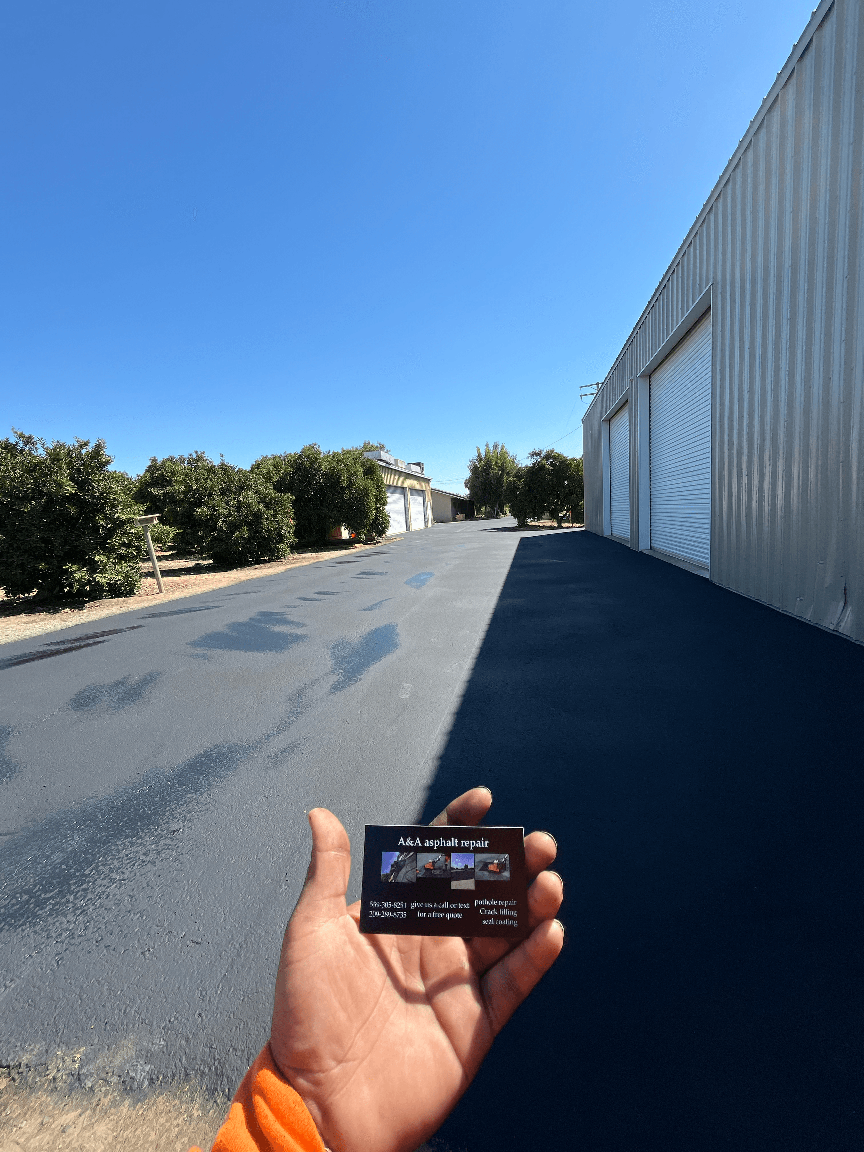Hand holding a business card for asphalt repair over a freshly sealed black asphalt driveway.